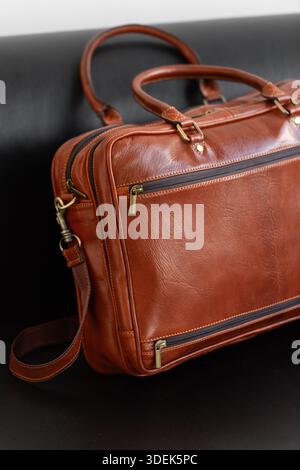 Une mallette professionnelle en cuir cognac avec deux poignées et lanière réglable se trouve sur un élégant canapé en cuir noir dans un cadre de studio minimaliste Banque D'Images