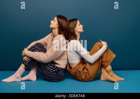 Deux soeurs partagent un moment de connexion, assis dos à dos dans des tenues chaudes et élégantes. Banque D'Images