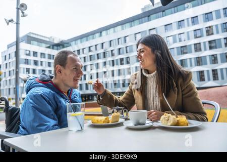 Femme nourrissant un homme souriant avec un handicap, profitant d'un repas et d'un café dans un café en plein air, incarnant des moments de soins, de compagnie et d'enj partagé Banque D'Images