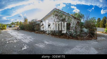 Une maison abandonnée en bois blanc de style méditerranéen, couverte de vignes vertes denses et de lierre, se dresse sur un terrain de gravier sous un ciel bleu vif Banque D'Images