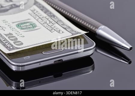 Nouveau smartphone, stylo à bille et cent dollars couchés sur une surface réfléchissante noire Banque D'Images