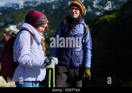 Deux randonneurs se tiennent debout sur une montagne enneigée, vêtus de vestes gonflantes, de chapeaux en tricot et de gants. Ils portent des sacs à dos et des bâtons de trekking, partageant un moment Banque D'Images