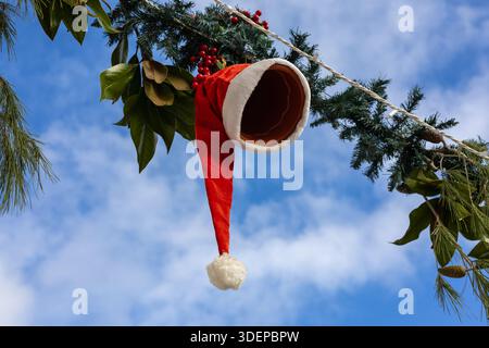 description décoration créative de noël avec un chapeau de père noël placé sur un pot en terre cuite suspendu à une branche de pin avec des baies rouges contre un bleu Banque D'Images