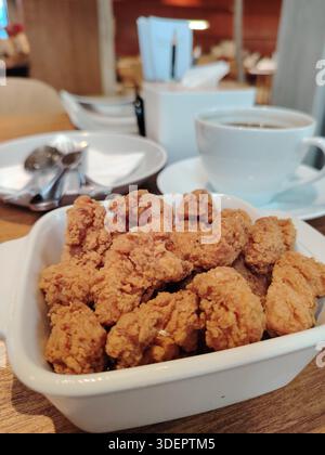 Poulet popcorn frit croquant dans un bol blanc, servi sur une table de café avec un fond de tasse de café flou. Banque D'Images