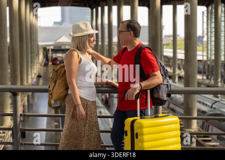 Heureux couple de touristes matures debout à l'intérieur d'une grande gare avec l'architecture moderne et les trains ci-dessous. Ils sourient et apprécient leur jou Banque D'Images
