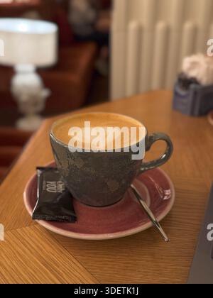 Cappuccino frais dans une tasse en céramique servi avec une soucoupe et une cuillère, placé sur une table de café en bois confortable. Banque D'Images