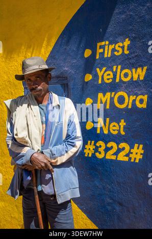 Un malgache vêtu d'une tenue distinctive, avec un chapeau et un châle de lamba drapé sur son épaule, s'appuie sur sa canne de marche tout en posant devant un mur de maison peint de couleurs vives. Camp Robin, Madagascar. Camp Robin est une commune rurale des hauts plateaux du centre de Madagascar. Il appartient au district d'Ambohimahasoa, qui fait partie de la région de haute Matsiatra Banque D'Images