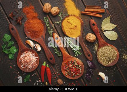 Différents types d'épices sont disposés dans des cuillères en bois sur une table sombre. Il y a des herbes, des graines et des feuilles placées à côté des cuillères créant une couleur Banque D'Images