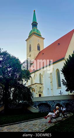 Une église historique dotée d'une grande flèche verte et d'un toit en tuiles rouges se trouve à côté d'un mur de pierre et d'un chemin pavé. Les visiteurs se reposent sur les bancs du parc, agréable Banque D'Images