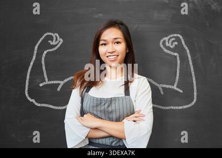Jeune femme asiatique en tant que fleuriste devant un tableau noir, fléchissant ses bras musclés, représentant un concept de vente au détail. Banque D'Images