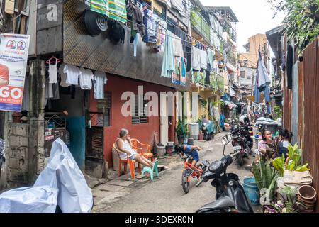 Vie quotidienne dans un bidonville urbain densément peuplé ou un établissement informel à Cebu, Philippines. La zone dispose de maisons de fortune et de ruelles étroites, où les résidents vivent à proximité. La photo montre des cordes à linge enfilées à travers les bâtiments, des gens assis à l'extérieur, illustrant l'activité communautaire dans des conditions exiguës. Banque D'Images