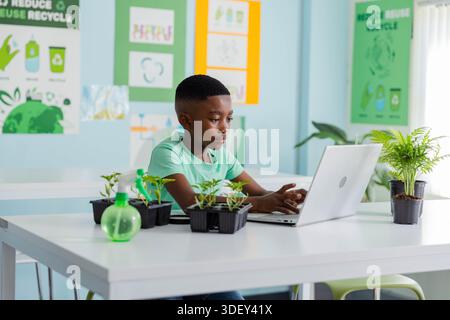 Étudiant afro-américain adolescent portant un tee-shirt vert travaillant sur ordinateur portable en classe avec des pots de plantes Banque D'Images