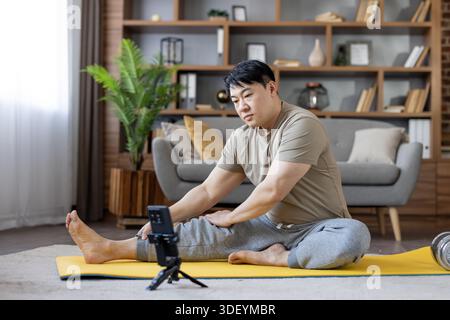 Homme asiatique sur un tapis de yoga jaune suivant une routine de fitness en ligne sur son smartphone à la maison, étirement et exercice dans le salon pour les wellnes Banque D'Images