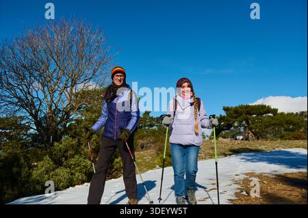 Deux randonneurs se tiennent sur un sentier enneigé sous un ciel bleu vif, souriant et habillé pour l’hiver. Ils tiennent des bâtons de trekking, prêts pour une journée d'avènement en plein air Banque D'Images
