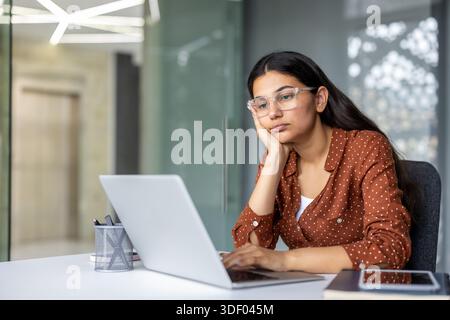 Jeune femme indienne éprouvant le stress au travail et l'ennui, reposant sa tête sur sa main tout en travaillant sur un ordinateur portable à un bureau moderne, se sentant épuisée Banque D'Images