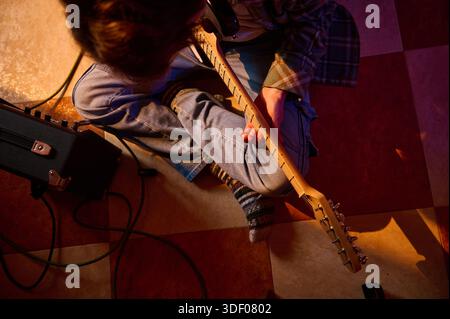Un musicien est assis sur le sol avec une guitare électrique, connectée à un amplificateur. Cadre chaleureux et intime avec chemise à carreaux, Jean en Jean et cabine enchevêtrée Banque D'Images