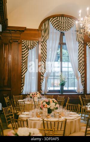 Les tables sont disposées avec des fleurs et de la vaisselle dans une pièce bien éclairée avec de grandes fenêtres et des accents en bois pour un rassemblement. Banque D'Images
