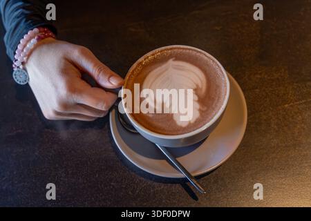 Main tenant une tasse de chocolat chaud avec latte La main d'une personne portant des bracelets perlés tient une tasse blanche de chocolat chaud avec latte en forme de feuille Banque D'Images