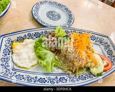 Assiette de riz frit avec de la viande râpée, des légumes et des oeufs frits servis sur un plat à motifs avec des concombres et des tranches de tomates - nasi goreng malaisien Banque D'Images