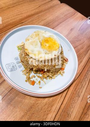 Assiette de nasi goreng, qui est du riz frit de style malaisien indonésien avec des légumes mélangés et oeuf frit sur le dessus servi sur une table en bois dans un restaurant Banque D'Images
