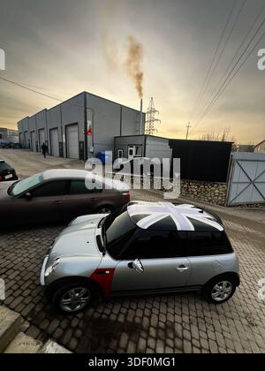 Lviv, Ukraine - 29 novembre 2025 : une Mini Cooper argentée avec un toit Union Jack garée sur des pavés dans une zone industrielle au coucher du soleil. Banque D'Images
