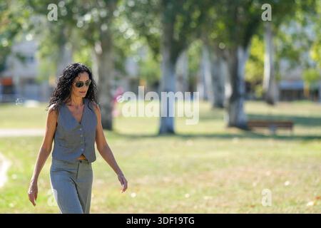 Une femme portant un gilet gris marche à travers un parc. Le parc est rempli d'arbres et de bancs Banque D'Images
