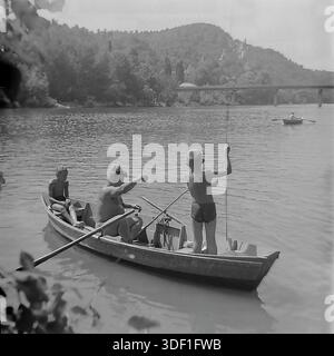 Sviatohirsk, région de Donetsk, Ukraine, URSS. 1970s. Un voyage de pêche en famille dans un bateau en bois sur la rivière Seversky Donets. Un homme adulte et deux garçons se détendent sur l'eau par une journée ensoleillée d'été. Un garçon est debout avec une canne à pêche. En arrière-plan, il y a un paysage pittoresque avec des montagnes de craie, une forêt dense et un pont en béton sur la rivière. Sur la colline, le monument à Artem et le complexe architectural du monastère sont visibles. Photo historique des vacances d'été en Union soviétique. Atmosphère authentique de la vie paisible, de l'enfance et des loisirs en plein air. Banque D'Images