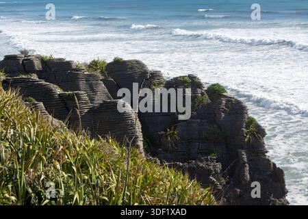 Une vue panoramique des formations rocheuses en couches le long d'une côte, avec des vagues s'écrasant contre le rivage. Une végétation verte luxuriante pousse sur les rochers, contrasti Banque D'Images
