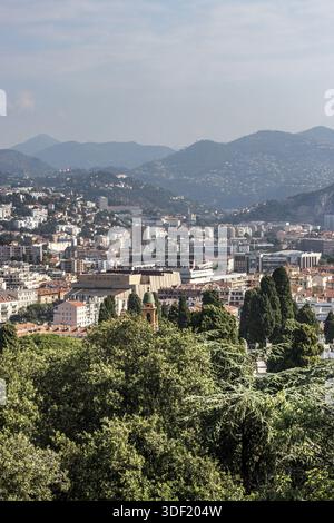 Vue sur les bâtiments baignés de soleil nichés parmi les arbres verdoyants, s'étendant vers les montagnes brumeuses lointaines sous un ciel clair, Nice, France. Banque D'Images