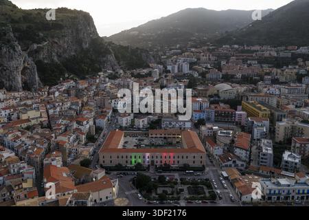 Vue aérienne des toits en terre cuite descendant en cascade pour rencontrer l'imposante structure illuminée de couleurs italiennes, encadrée par des montagnes escarpées, Cefalu, Banque D'Images