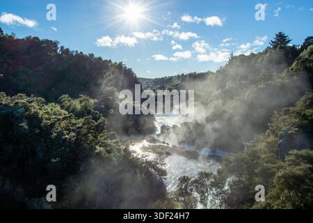 Un paysage serein avec une rivière qui coule à travers des collines verdoyantes, avec la brume qui monte de l'eau. Le soleil brille brillamment dans un ciel bleu clair, c Banque D'Images