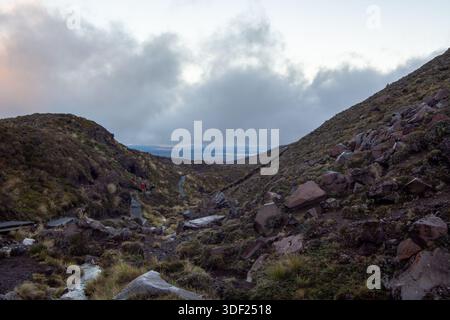 Une vue panoramique sur une vallée accidentée avec un terrain rocheux et des nuages bas. Deux randonneurs sont visibles sur un sentier sinueux, entouré d'une végétation luxuriante et de montagneuses Banque D'Images