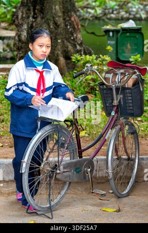 Écolière vietnamienne vérifiant ses devoirs sur son vélo, commune de SAI son, district de Quoc Oai, en périphérie de Hanoi, Vietnam Banque D'Images