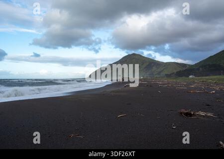 Une vue panoramique sur une plage de sable noir avec des vagues qui s'écrasent contre le rivage. En arrière-plan, une montagne verdoyante s'élève sous un ciel nuageux. Driftwoo Banque D'Images