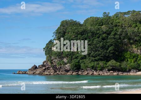 Une scène de plage sereine avec une côte rocheuse et une végétation luxuriante. Les eaux turquoises calmes tapent doucement contre le rivage, sous un ciel bleu clair d'esprit Banque D'Images