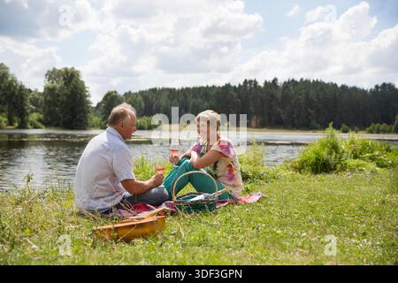 charmant couple de personnes âgées au lac ayant un pique-nique, faisant un toast sur la rive du lac un jour d'été. Staycations, voyage hyper-local, sortie en famille, escapade. || autorisation du modèle Banque D'Images