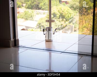 Pot de moka en métal sur un sol carrelé près d'une porte vitrée coulissante, représentant la préparation du café maison et la routine matinale. Banque D'Images