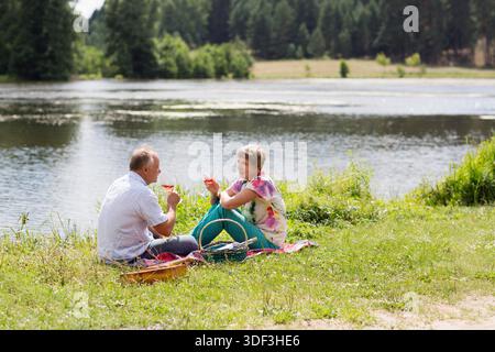 charmant couple de personnes âgées au lac ayant un pique-nique, faisant un toast sur la rive du lac un jour d'été. Staycations, voyage hyper-local, sortie en famille, escapade. || autorisation du modèle Banque D'Images