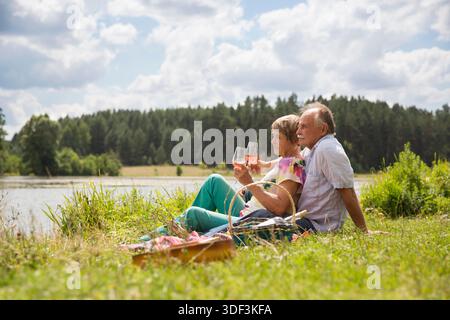 charmant couple de personnes âgées au lac ayant un pique-nique, faisant un toast sur la rive du lac un jour d'été. Staycations, voyage hyper-local, sortie en famille, escapade. || autorisation du modèle Banque D'Images
