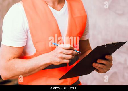 Ouvrier masculin en gilet orange vif utilisant un stylo pour écrire sur le presse-papiers dans un environnement intérieur flou Banque D'Images