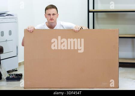 Homme tenant une grande feuille de carton dans le corps cachant avant. Homme adulte tenant un grand panneau de carton vierge devant lui-même debout dans une pièce minimaliste Banque D'Images