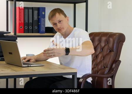 Bras extensible masculin occasionnel dans un geste explicatif tout en étant assis à un bureau en bois avec ordinateur portable ouvert et classeurs Banque D'Images