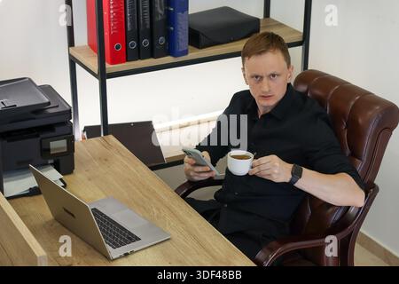 Professionnel irrité assis au bureau en bois avec boisson et appareil mobile, affichant le mécontentement dans le lieu de travail Banque D'Images