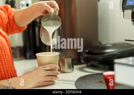 Vue rapprochée du barista versant du lait crémeux dans un gobelet en papier pour la préparation d'un latte dans un café professionnel. Banque D'Images
