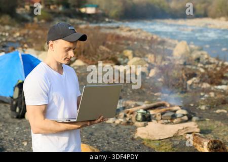 Un homme portant une casquette est assis avec un ordinateur portable ouvert près d'un feu de camp sur une rive rocheuse, tente dressée en arrière-plan sous le feuillage d'automne. Banque D'Images