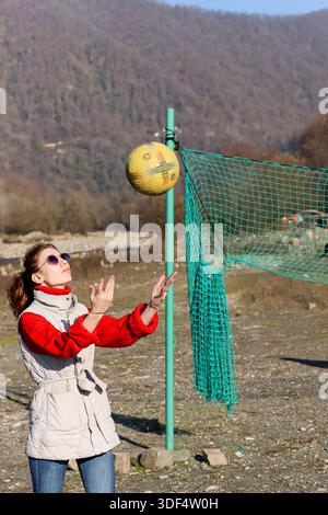 Femme dans des vêtements en couches atteint pour descendre le ballon de football à côté du poteau de filet de fortune dans un environnement extérieur accidenté avec des collines. Banque D'Images