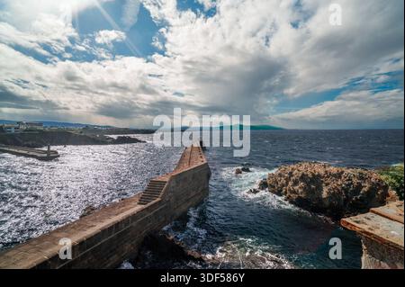Jetée en pierre s'étendant dans les eaux agitées de la mer Cantabrique, avec l'architecture de la ville côtière et les collines verdoyantes lointaines sous un ciel lumineux et nuageux dedans Banque D'Images