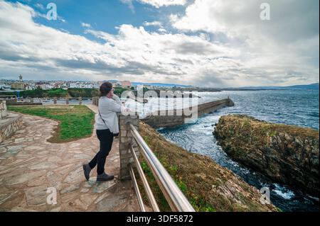 Femme debout sur un point de vue côtier, regardant le rivage accidenté, un long brise-lames protégeant le port de pêche de Tapia de Casariego, AST Banque D'Images