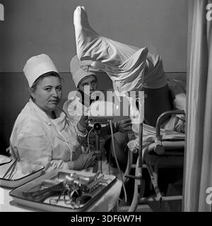 Slaviansk, RSS d'Ukraine, années 1970 Un cliché historique représente un examen gynécologique dans un hôpital soviétique. Deux femmes médecins en blouses et casquettes de coton blanc s'occupent d'une patiente sur une chaise gynécologique. Le médecin principal regarde la caméra, tandis que l'assistant ajuste un colposcope, un instrument optique spécialisé pour le diagnostic médical. Un plateau avec des instruments en métal stérilisés se trouve au premier plan. Cette authentique photographie d'archives monochrome illustre l'accent mis sur la santé des femmes et les soins médicaux professionnels dans la paisible région du Donbass. Banque D'Images