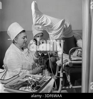 Slaviansk, RSS d'Ukraine, années 1970 Un cliché historique capture un bilan gynécologique dans une clinique soviétique. Une femme médecin senior dans une robe de coton blanche utilise un colposcope pour des diagnostics détaillés, en regardant dans les oculaires. Une jeune infirmière assiste à proximité. Le patient est placé sur une chaise recouverte d'un drap. Un plateau avec des spéculums métalliques se trouve au premier plan. Cette authentique photographie d'archives monochrome reflète les normes élevées des soins de santé des femmes et la routine médicale quotidienne dans la paisible région du Donbass. Banque D'Images